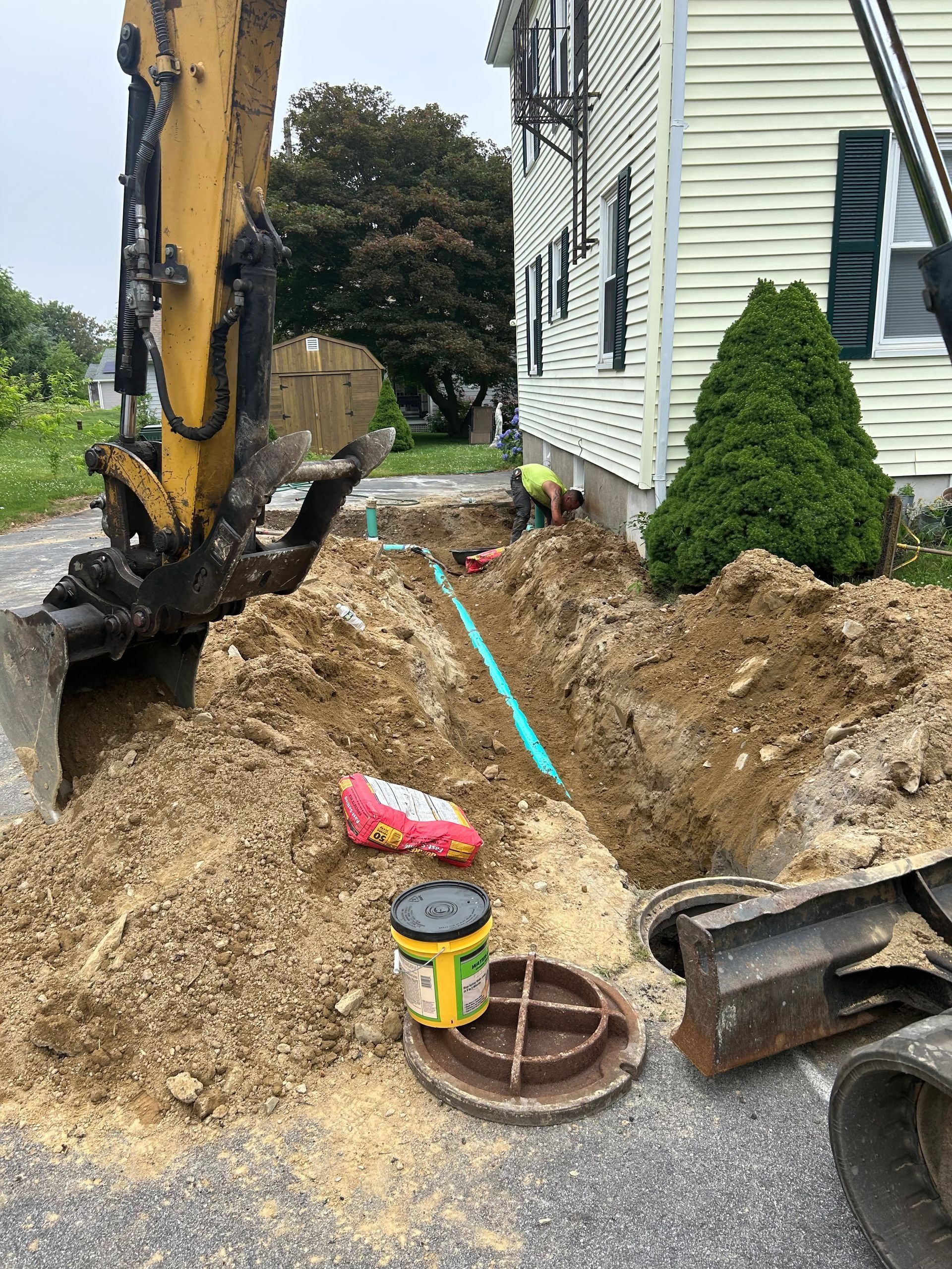 An excavator digs a trench next to a house where a worker installs a green sewer pipe near a utility access cover.
