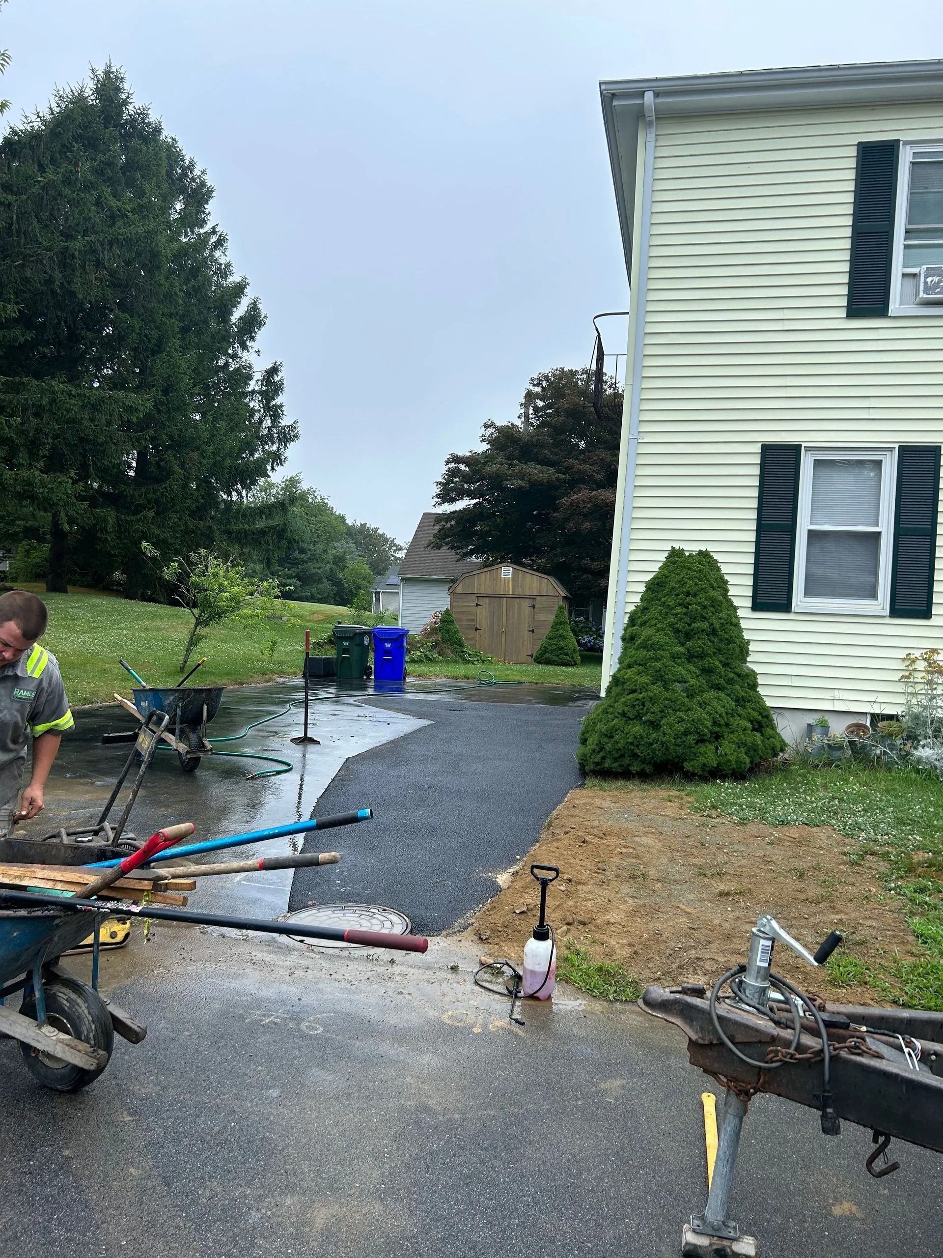 A worker in a high-visibility vest stands near a wheelbarrow and driveway construction next to a light yellow house.