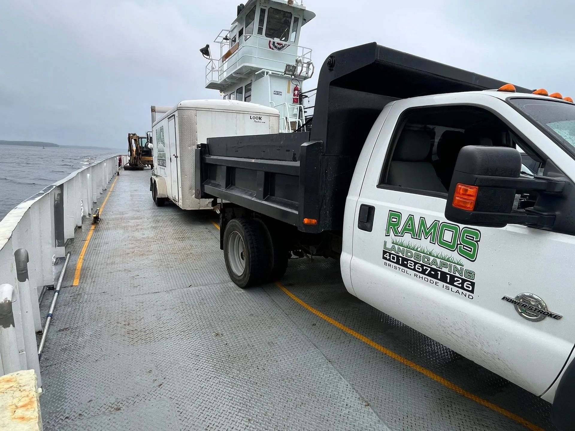 A white dump truck pulling a trailer on the metal deck of a ferry crossing a body of water.