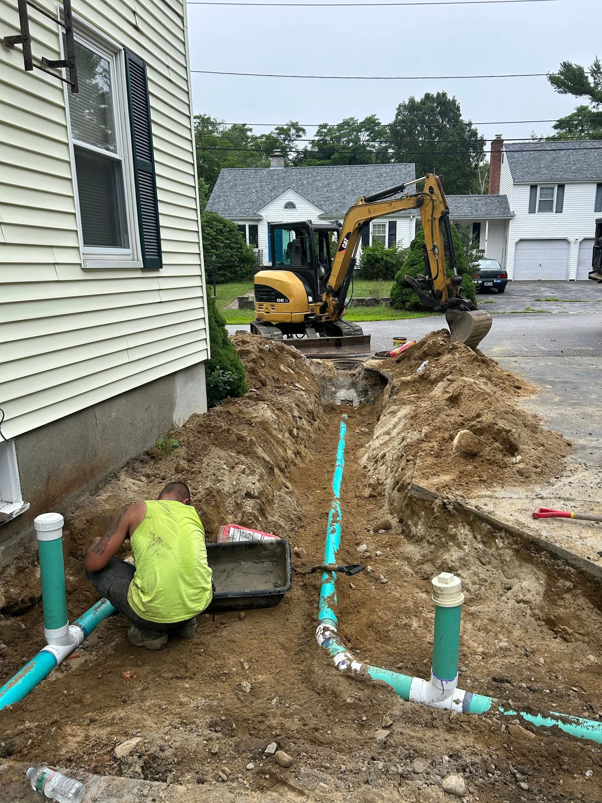 A construction worker in a neon vest installs green pipes in a trench next to a house with a yellow excavator in the yard.