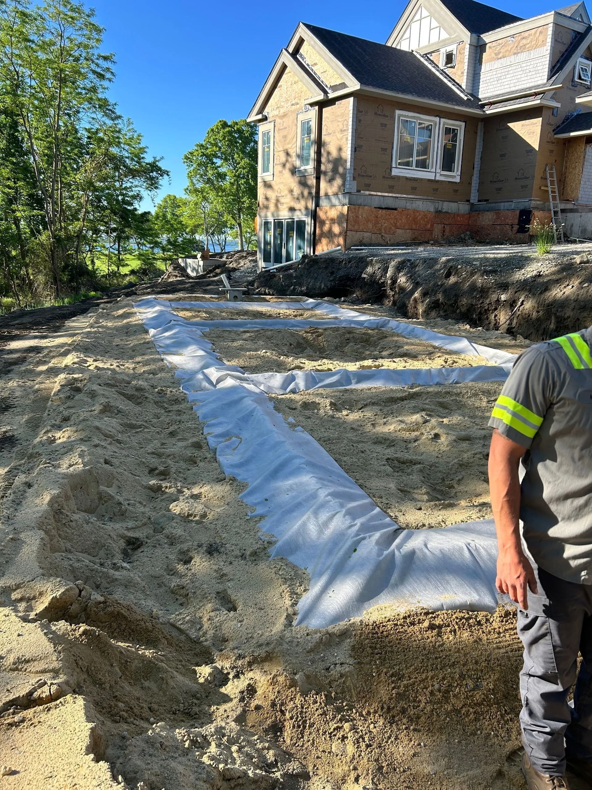 A worker stands near a trench lined with white geotextile fabric at a residential construction site.