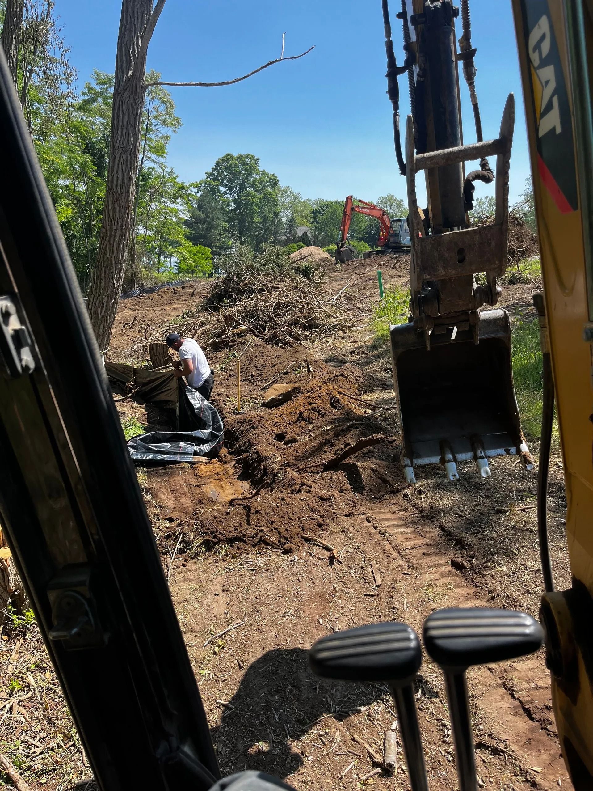 View from the cab of a CAT excavator showing a worker digging in a wooded, cleared construction site.