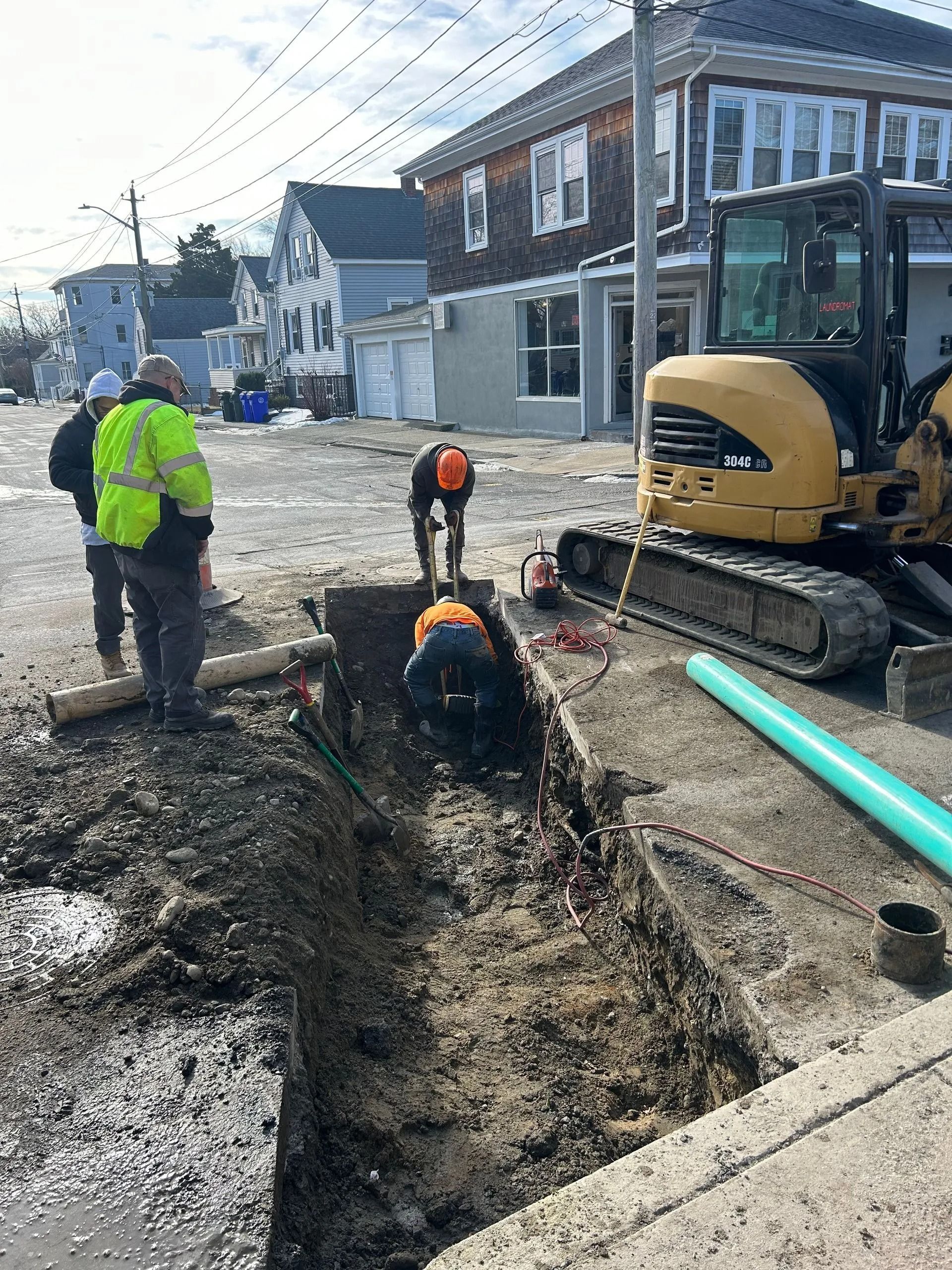 Construction workers install a green pipe in a street-level trench next to a yellow excavator.
