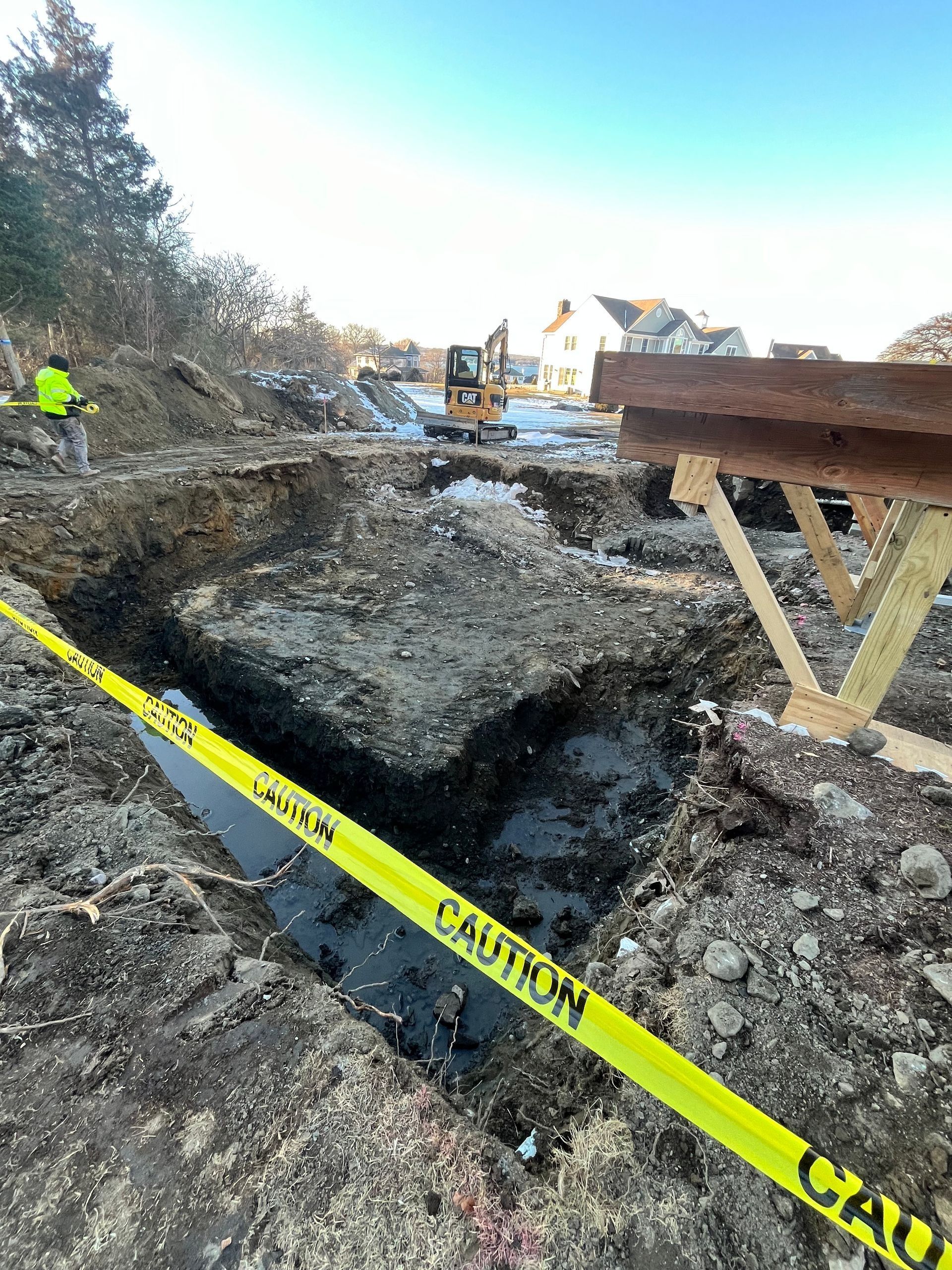 An excavation site featuring a muddy trench, a worker in a high-visibility vest, and yellow caution tape in the foreground.