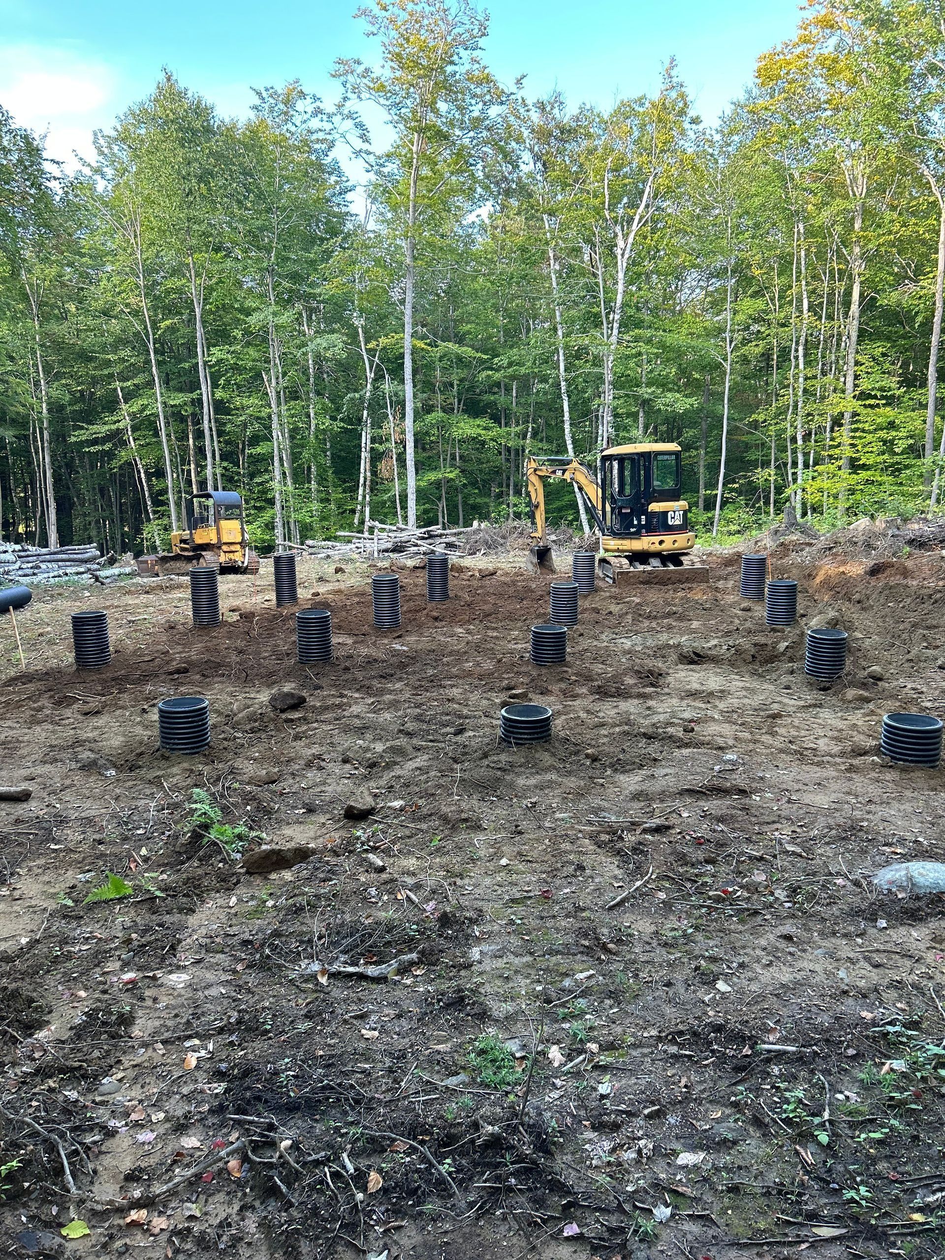 Construction site in a wooded area showing rows of black cylindrical foundation forms and yellow excavating machinery.