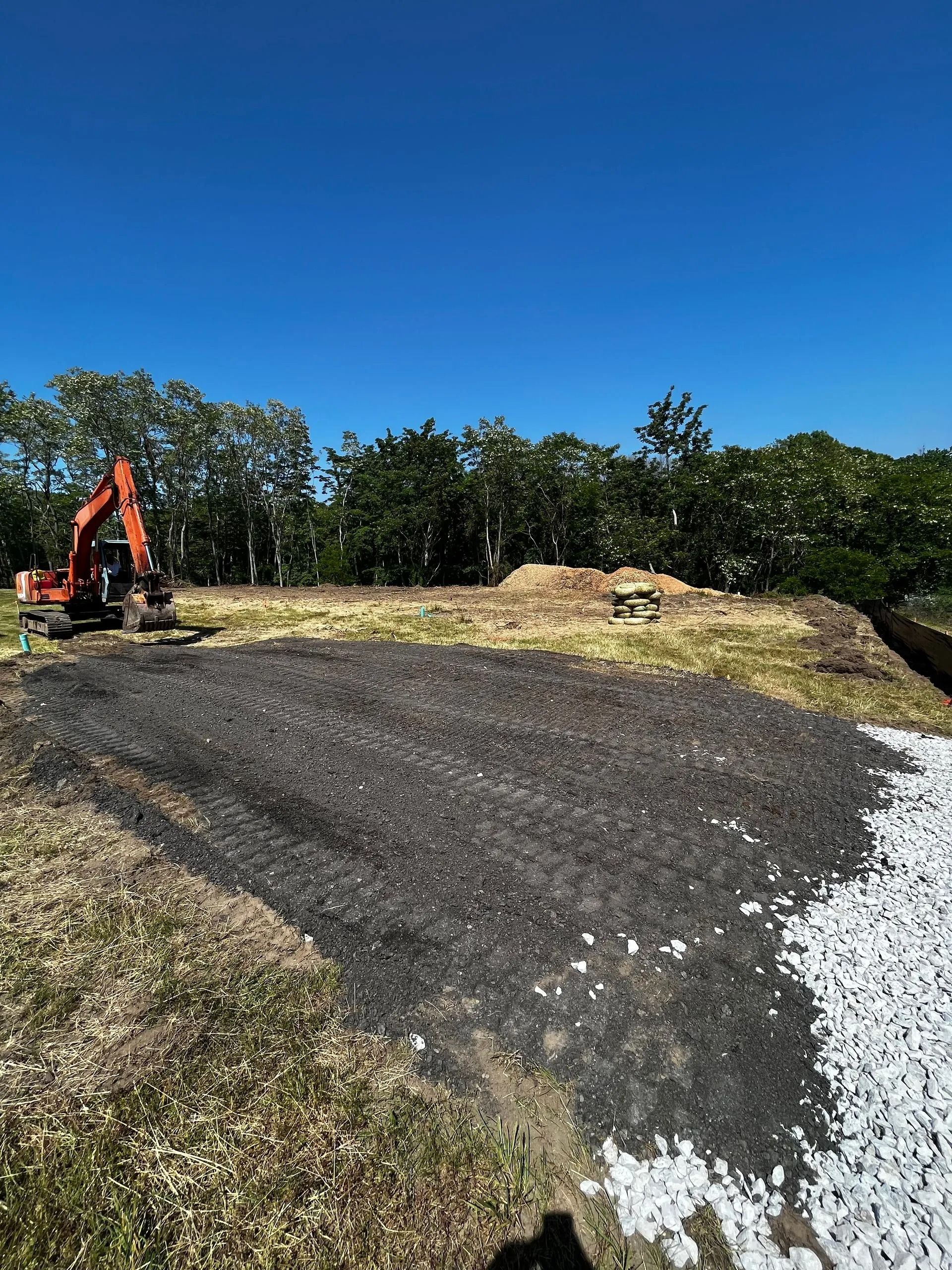 An orange excavator sits on a grassy construction site with dark gravel and white rocks spread across the ground.