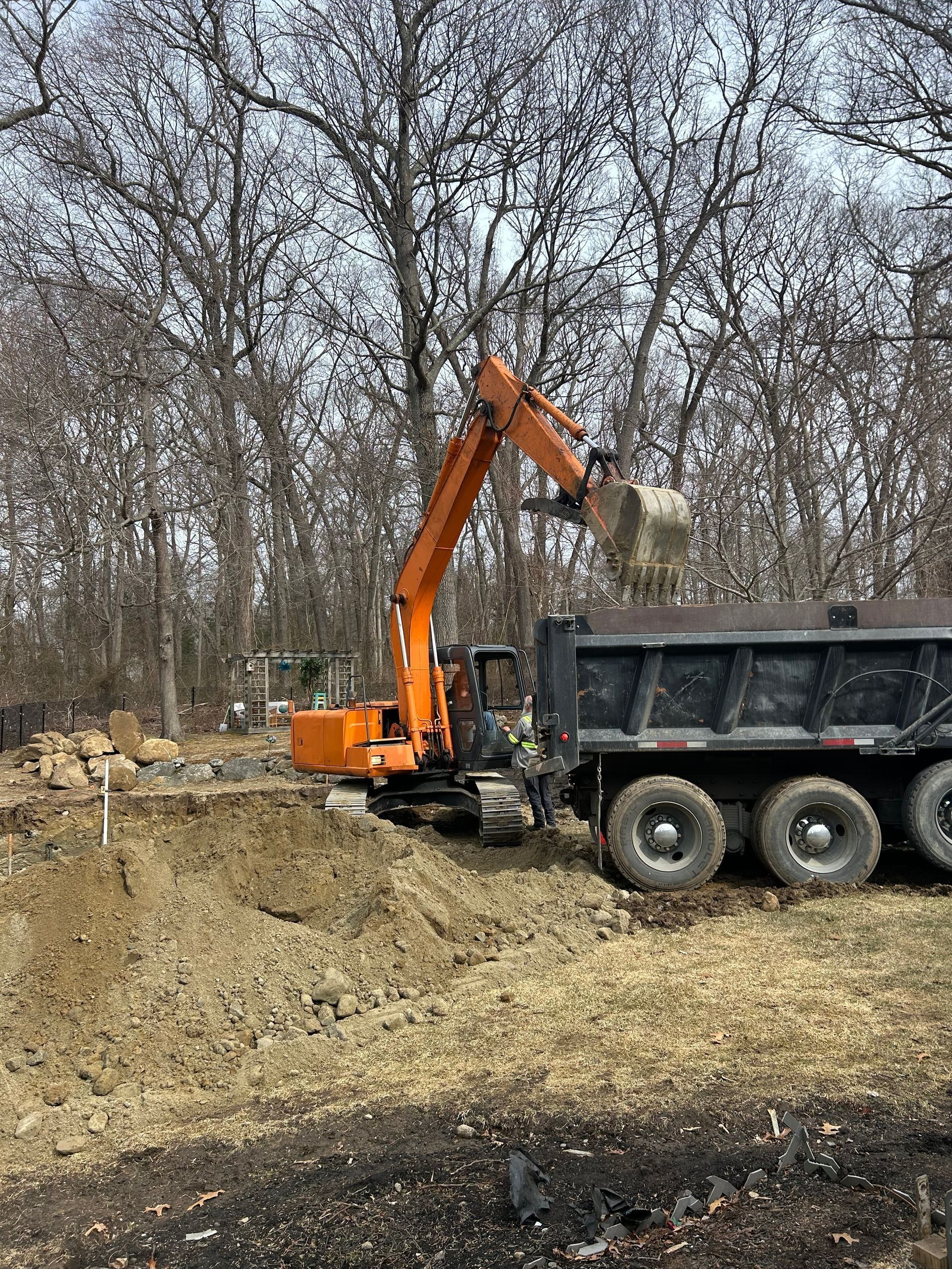 An orange excavator fills a dump truck with soil at a wooded construction site.