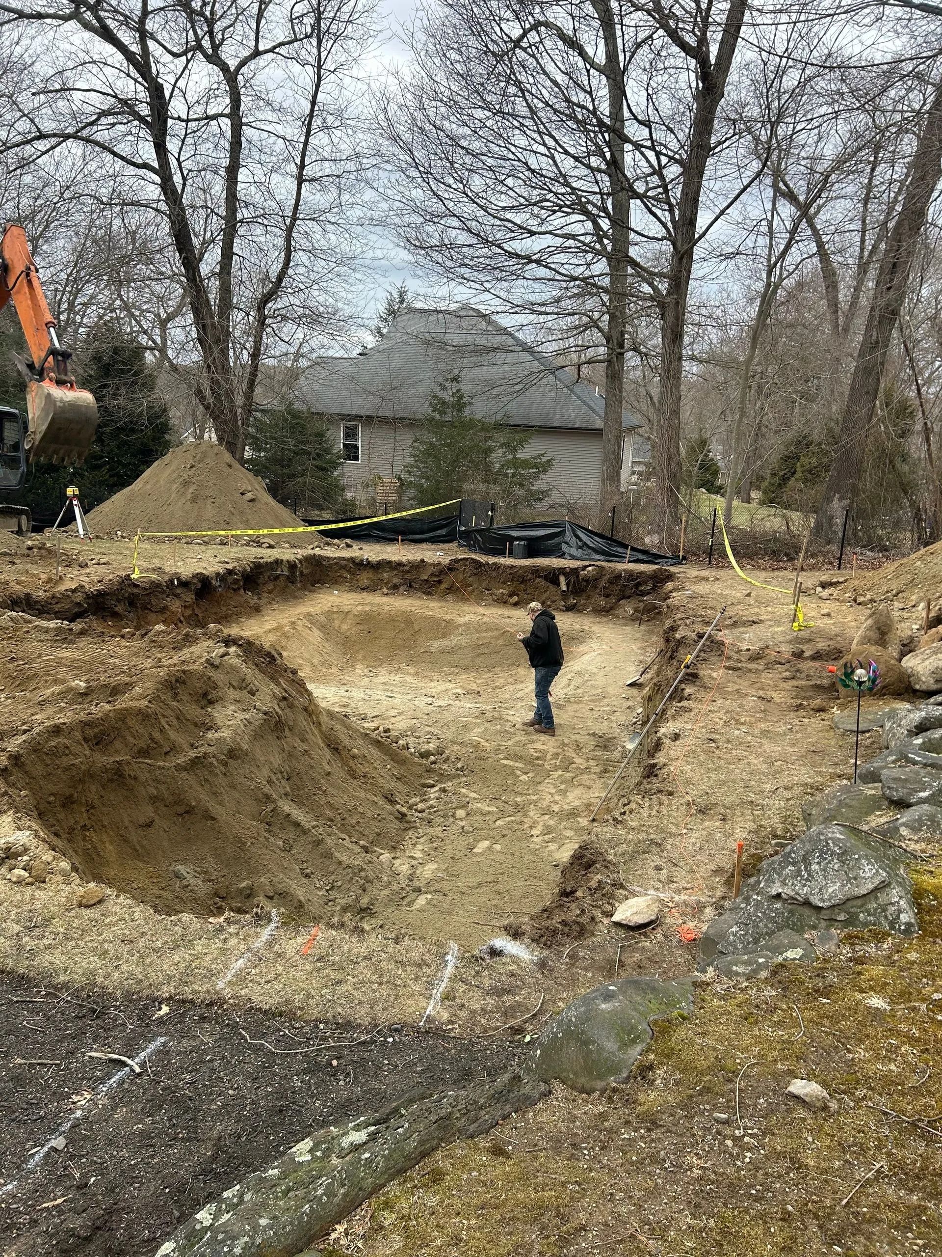 A person stands in a large, excavated dirt pit for a construction project, with a house and bare trees in the background.