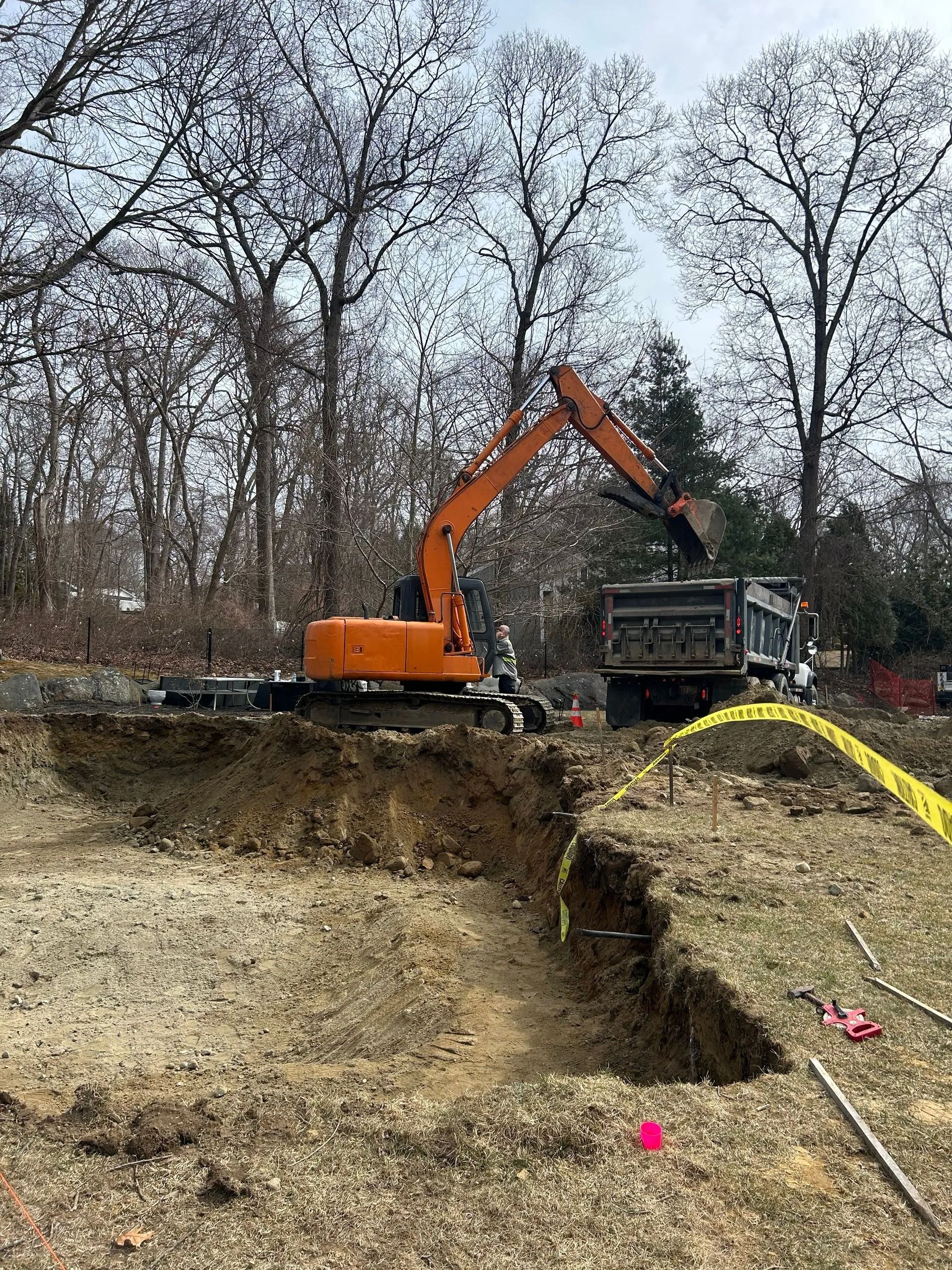 An orange excavator sits on a dirt mound, loading soil into a dump truck at a construction site with trees in the back.