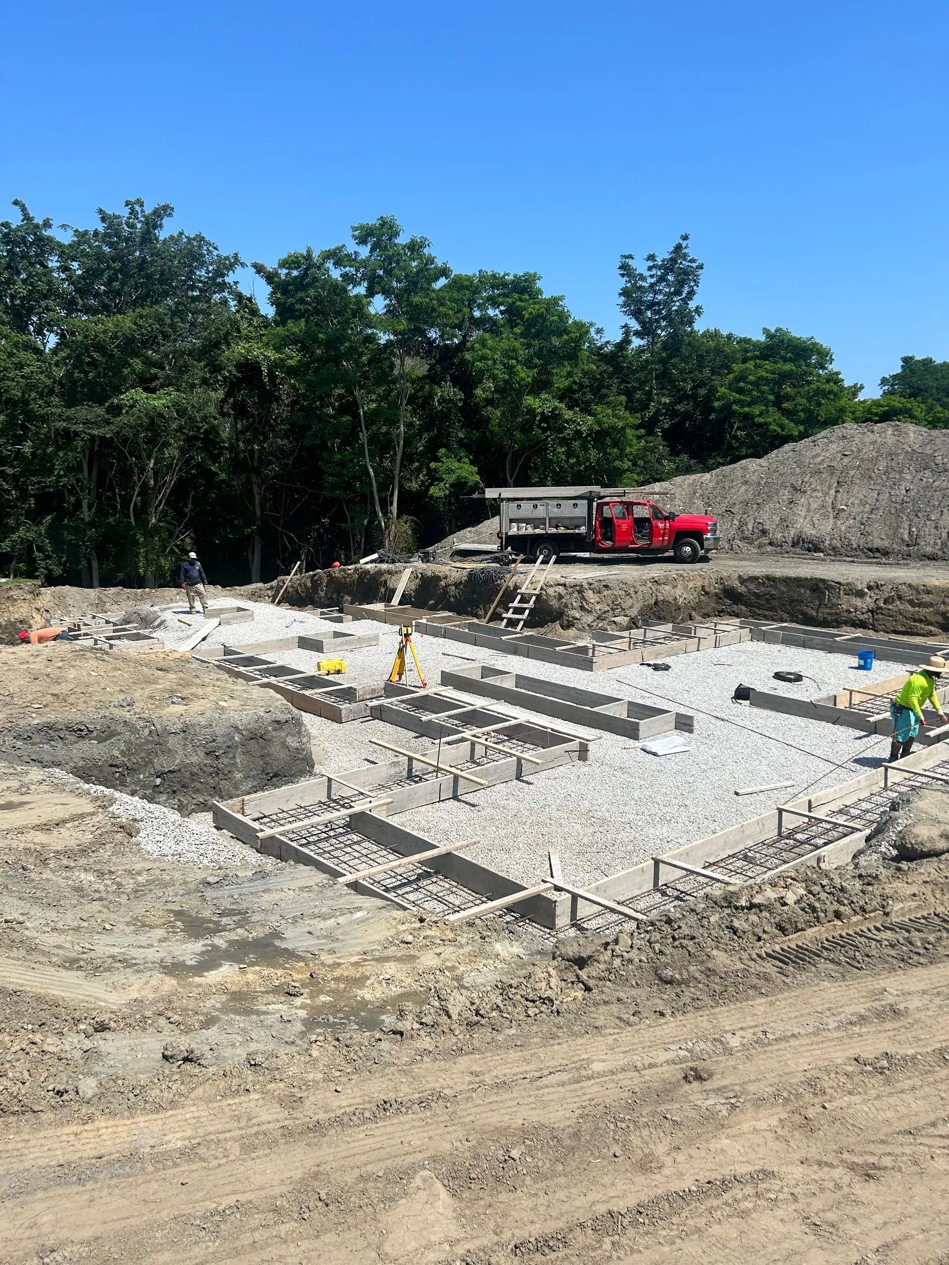A red dump truck parked at a construction site with workers laying concrete blocks on a gravel foundation.