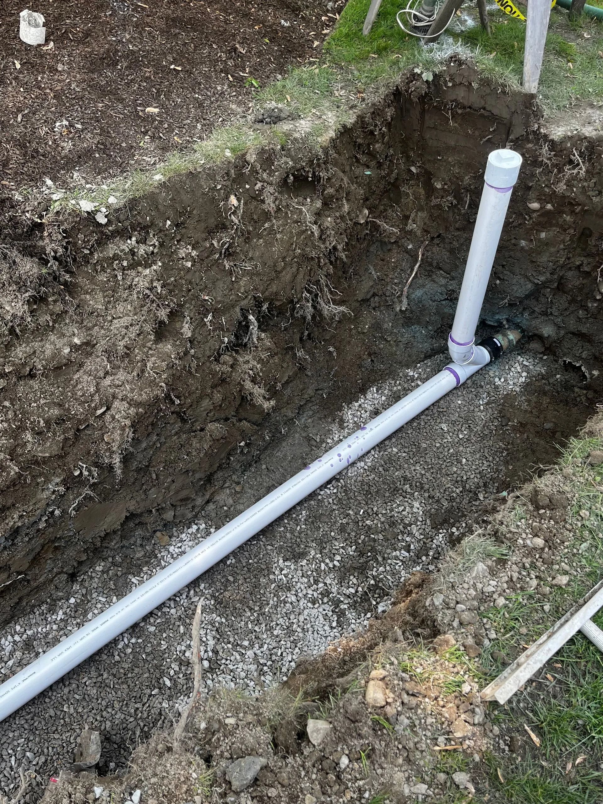 A white PVC pipe installed in a gravel-lined trench with a vertical pipe extending upward.