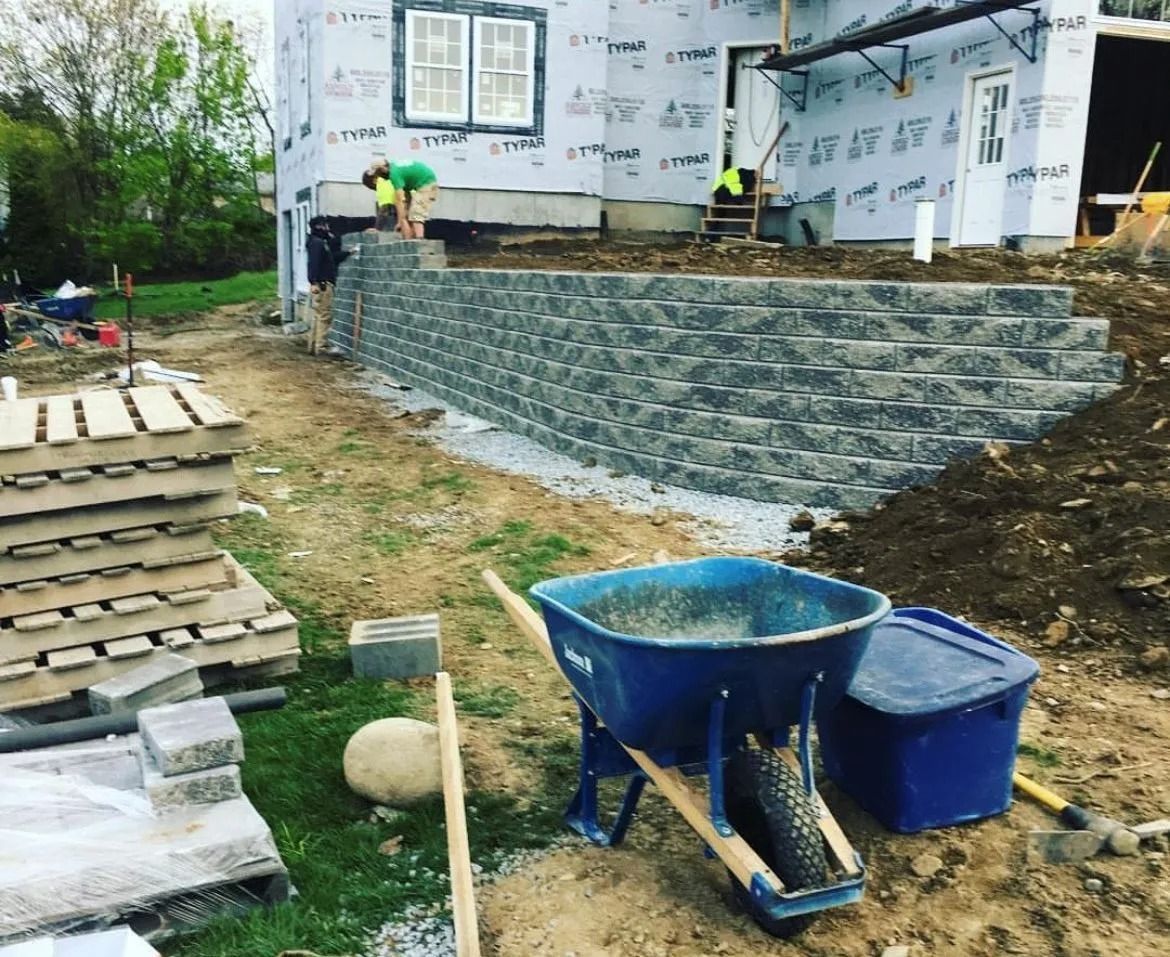Construction workers build a gray stone retaining wall in front of a house under renovation near a blue wheelbarrow.