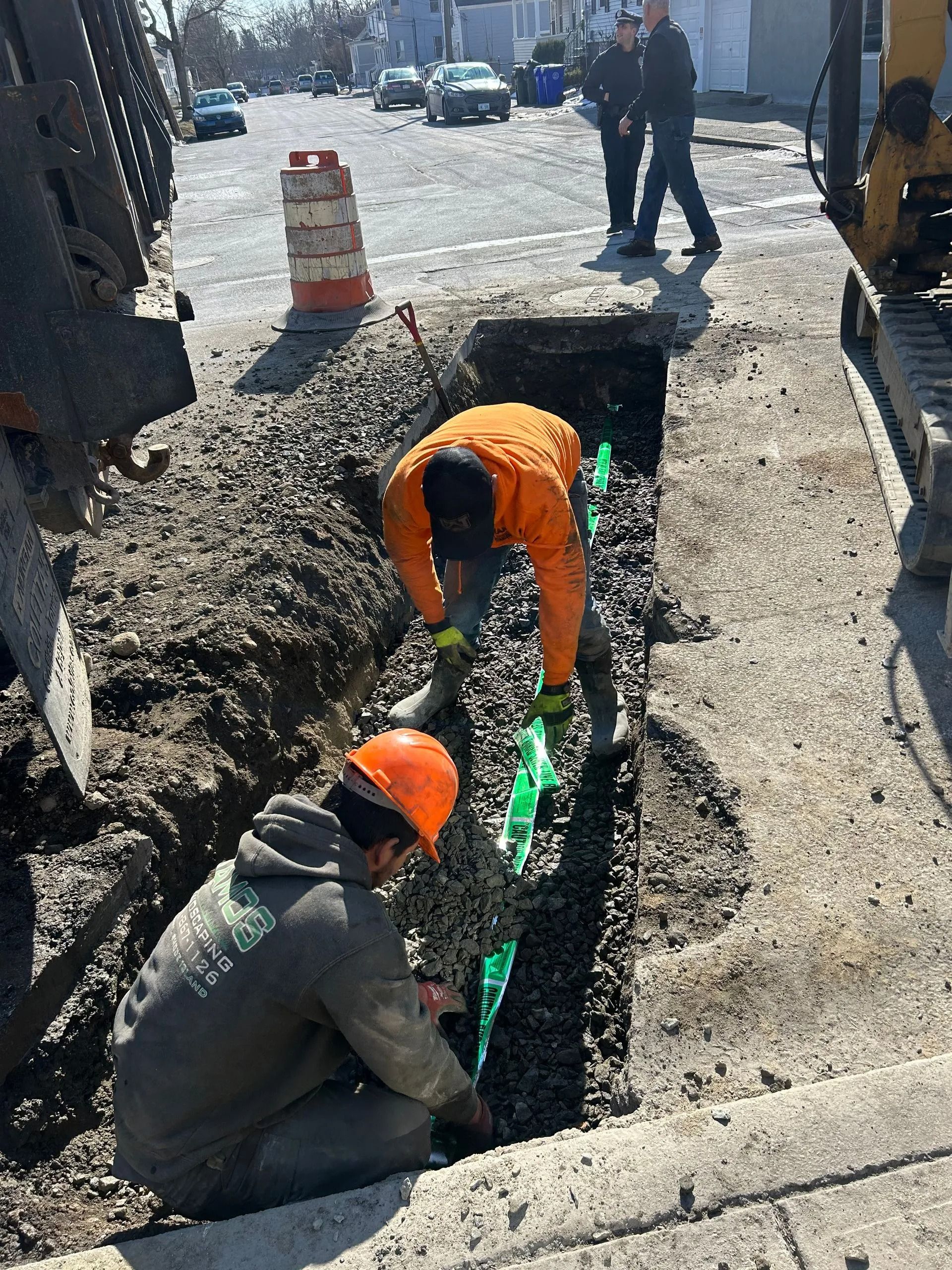 Two workers in safety gear install a utility line in a narrow trench on a sunny street.
