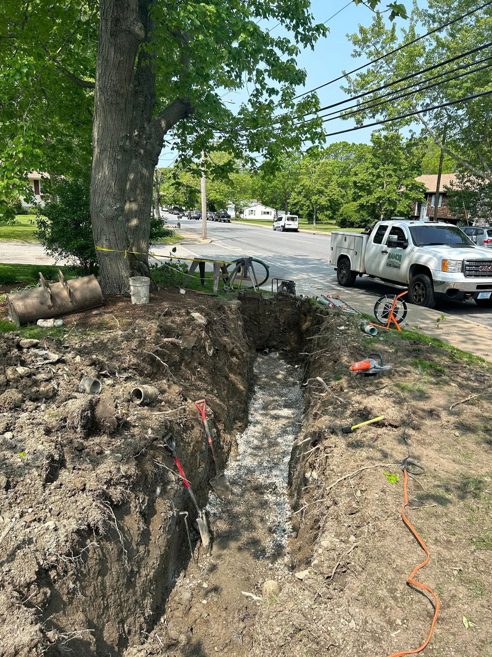 A deep, narrow trench dug into the ground next to a tree and a utility truck parked on a sunny residential street.