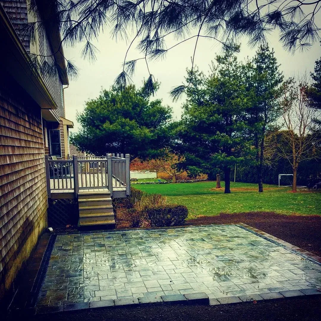 A newly laid stone patio next to a wooden deck attached to the side of a house, overlooking a grassy backyard.