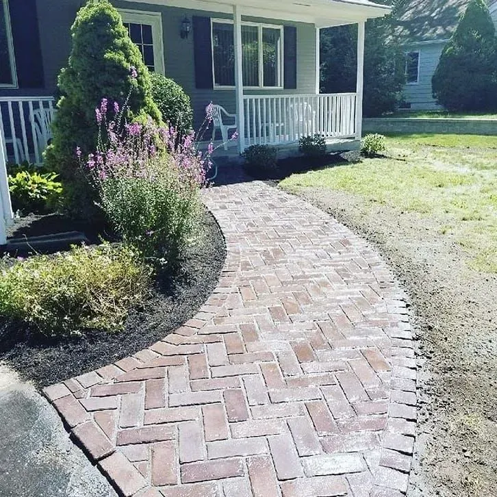 A curved herringbone brick walkway leads to the front porch of a gray house surrounded by landscaping.