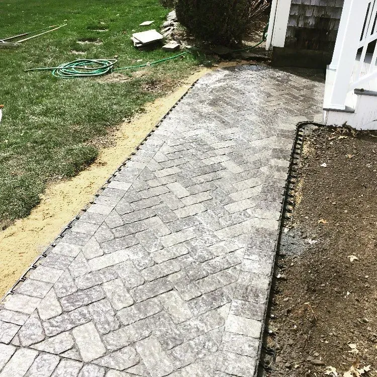 A new herringbone-pattern paver walkway runs along the side of a house, flanked by a dirt area and a green lawn.