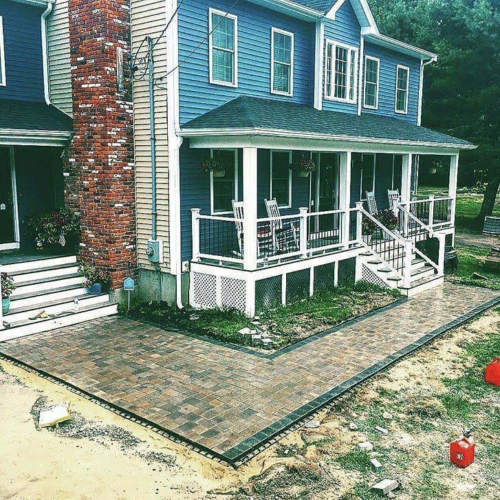 A blue, two-story house with a brick chimney, a front porch with white railings, and a newly installed paved patio.