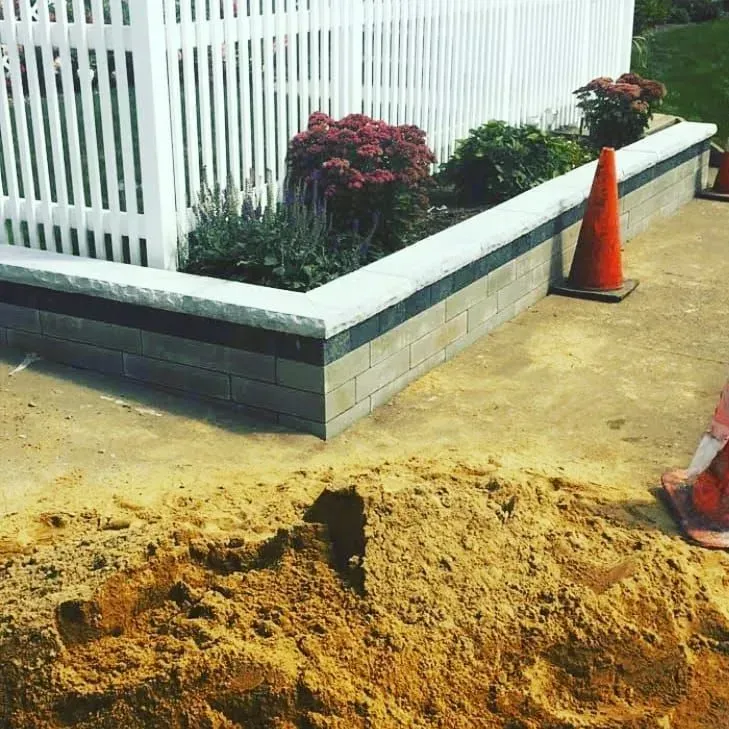 A low stone retaining wall with a white picket fence, green shrubs, and a pile of sand in the foreground.