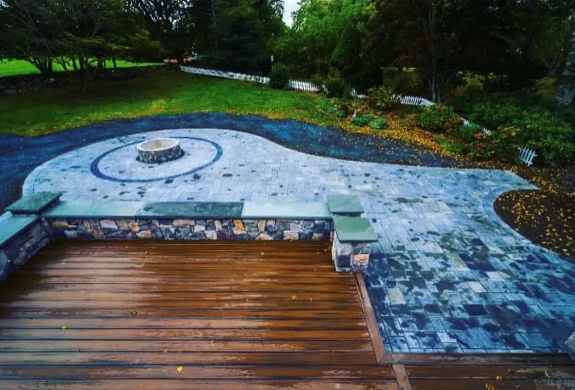A backyard view showing a wooden deck transitioning into a curved, multi-toned stone patio with a central circular fire pit.