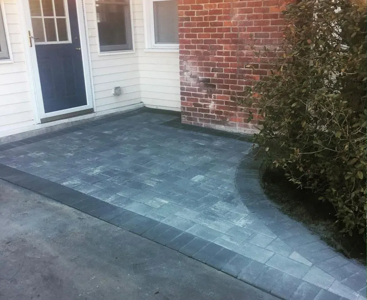 A new stone patio featuring dark grey pavers with a border, adjacent to a white house with a brick chimney.