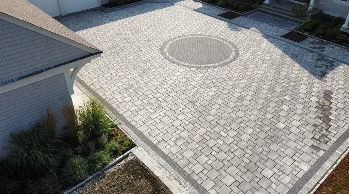 An aerial view of a gray paver driveway with a circular pattern centerpiece, bordered by a house and landscaping.