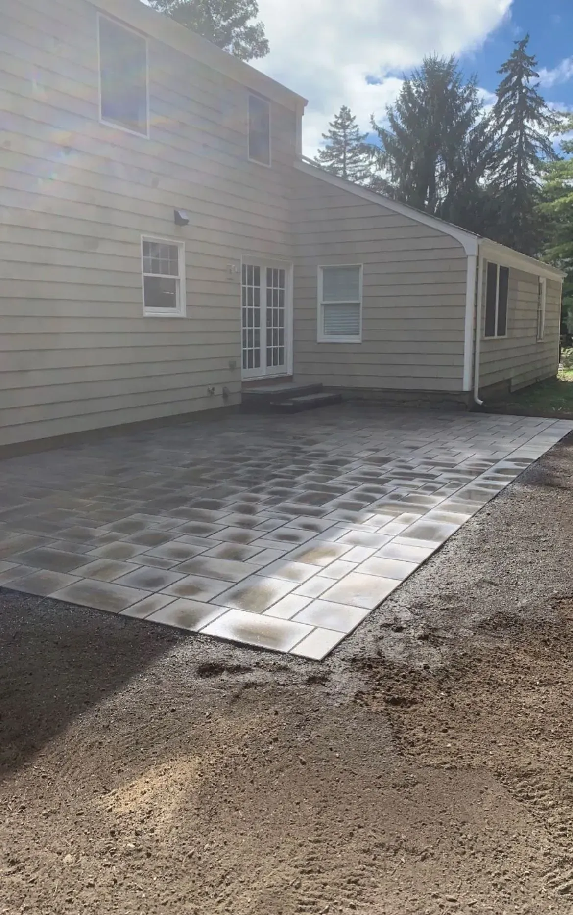 A new gray and tan paver patio installed against the side of a light-colored, two-story house with a lawn area.