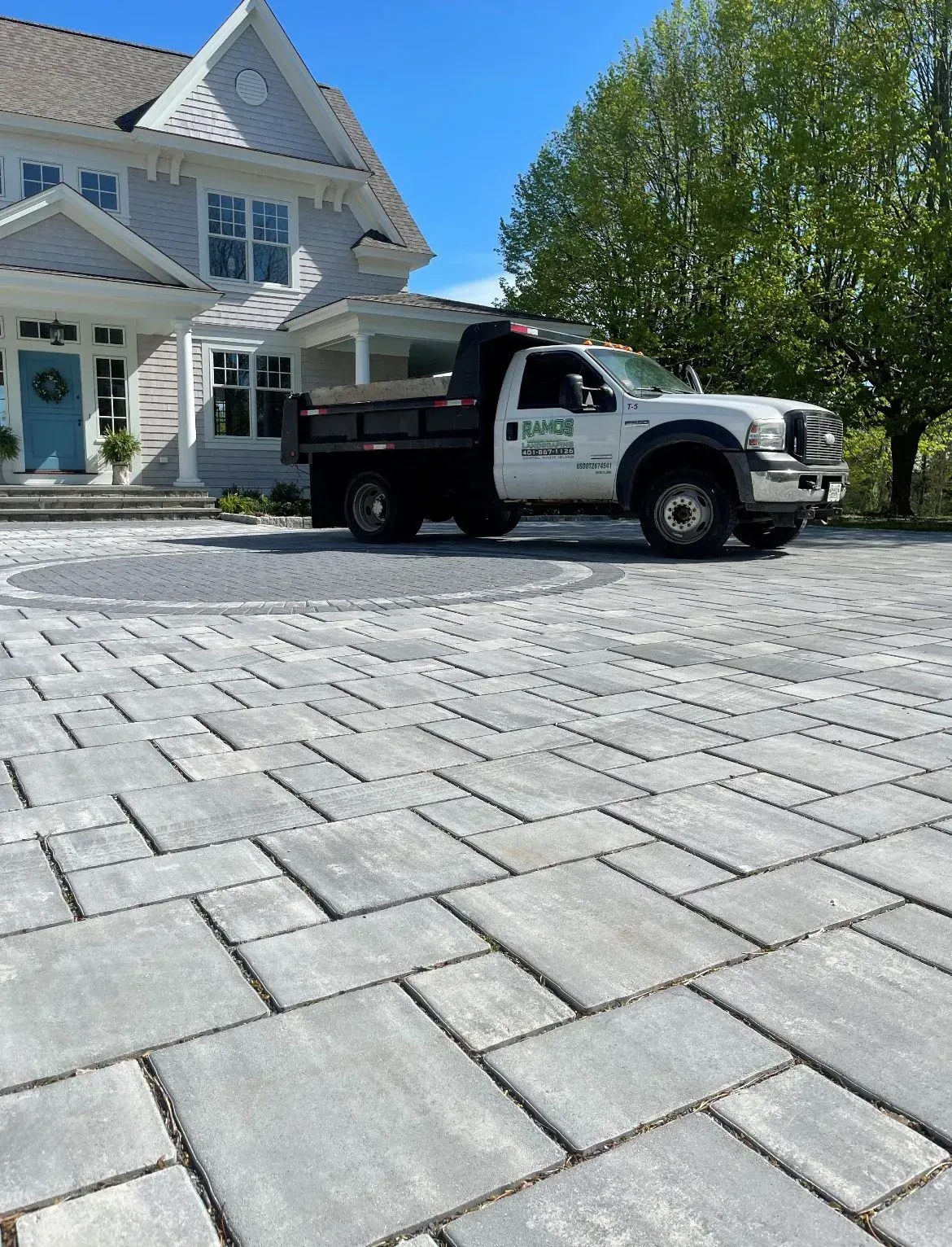 A white dump truck with a company logo parked on a large, light-gray stone paver driveway in front of a modern house.