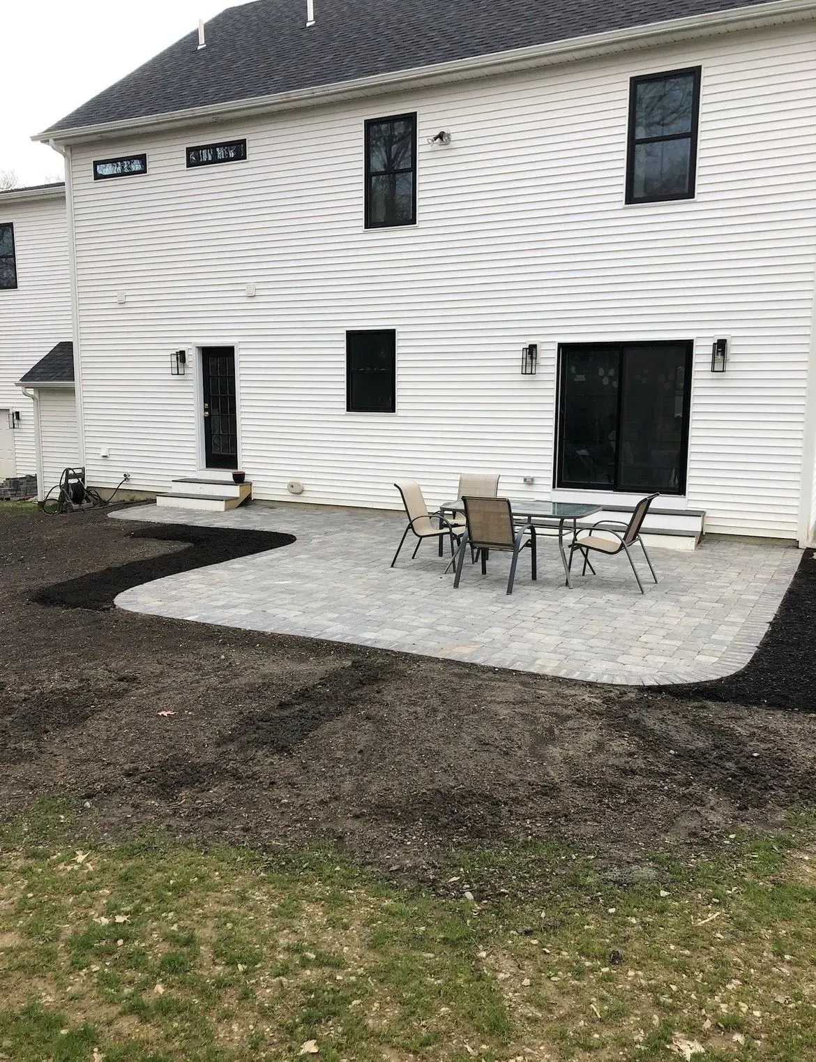 A gray stone patio with a patio table and chairs sits behind a white house with black trim and windows.