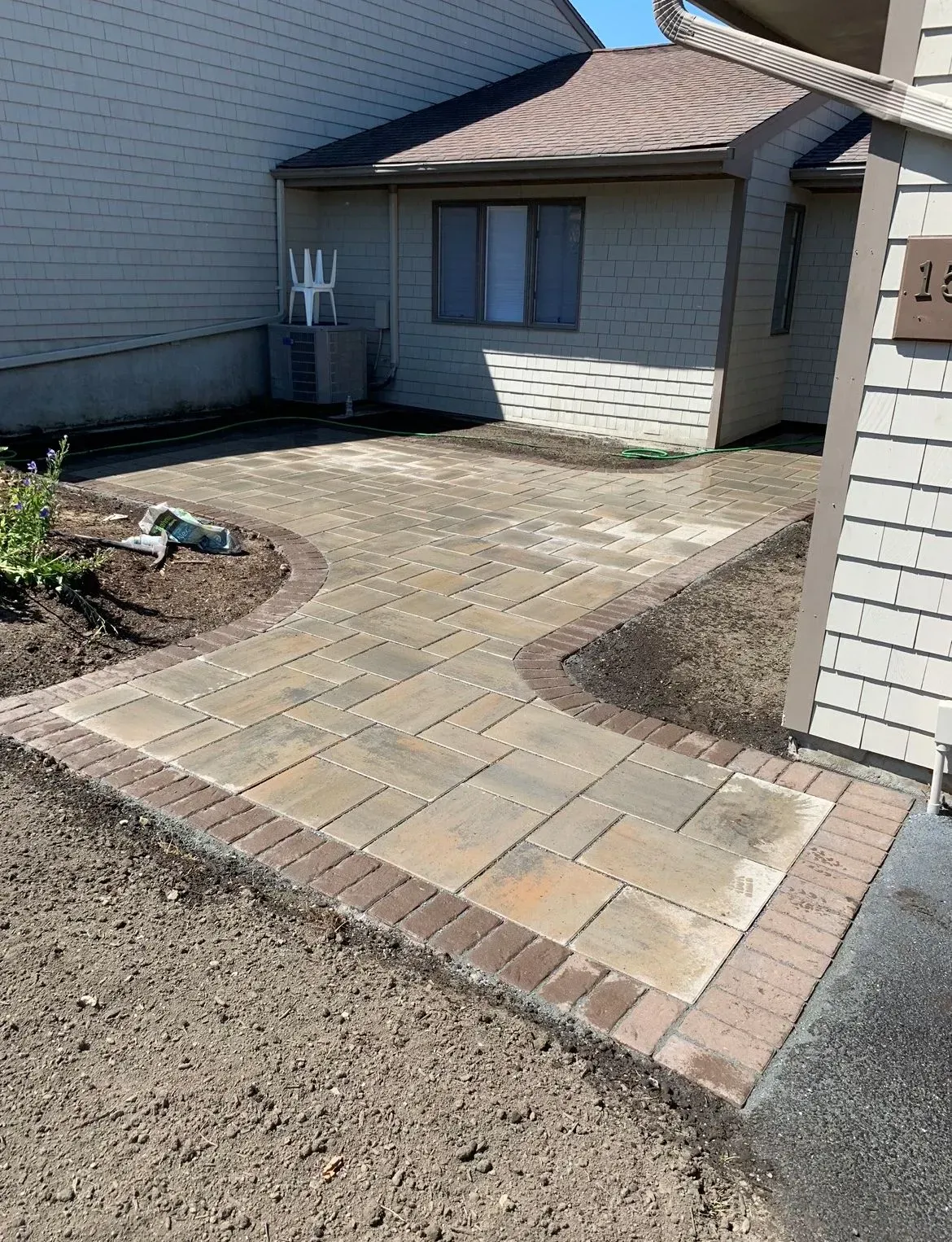 A newly installed paver walkway leads to the entrance of a house, surrounded by bare soil and exterior siding.
