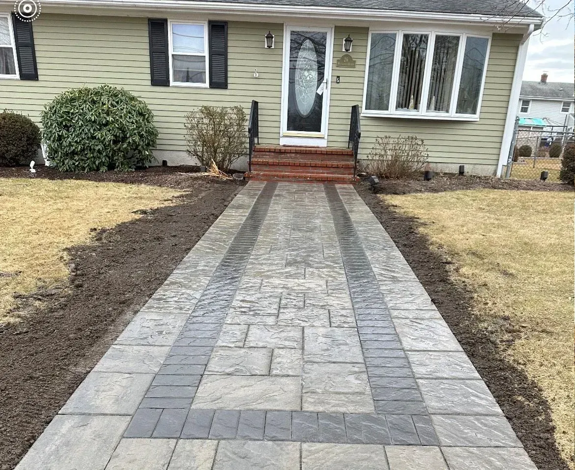 A stone walkway with a dark grey border leads to the front entrance of a house with pale green siding.