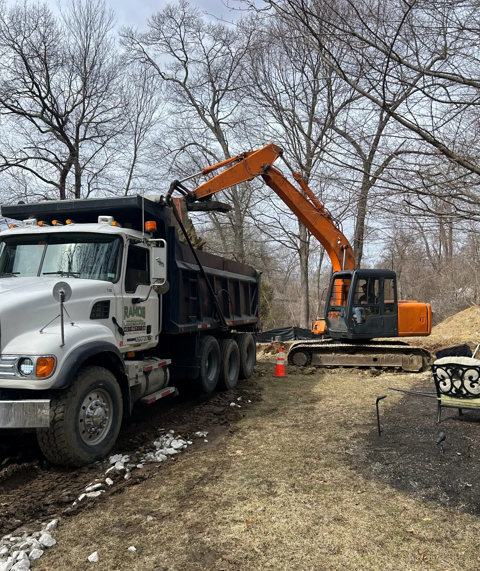 An orange excavator loads dirt into the back of a white dump truck parked on a dirt path near bare trees.