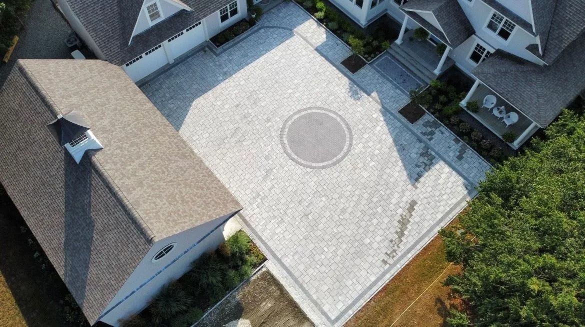 An aerial view of a large residential driveway paved with light gray stone, featuring a circular inlay in the center.
