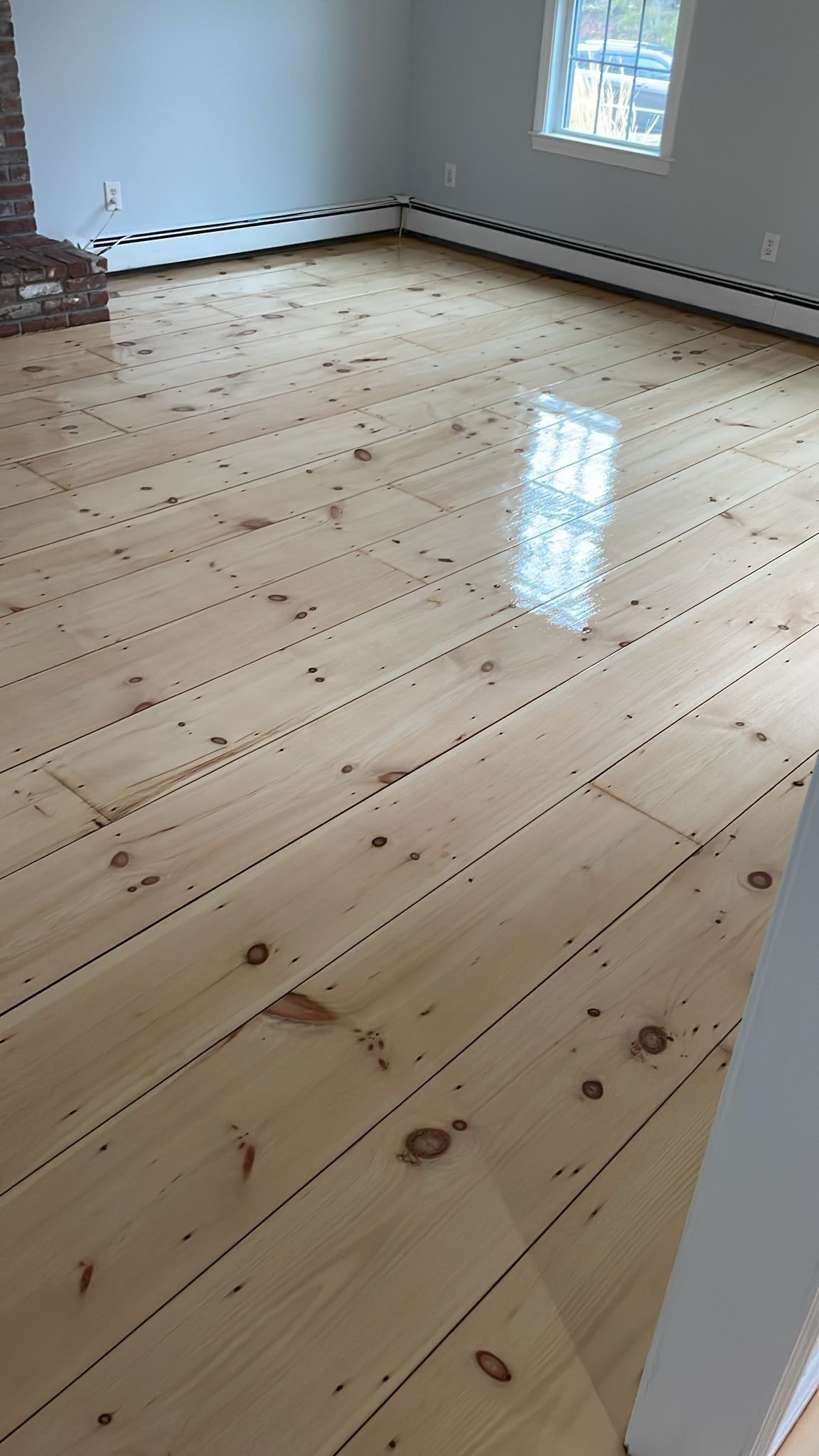 Light-colored wooden floor in a room with a window, baseboard heaters, and a brick fireplace.