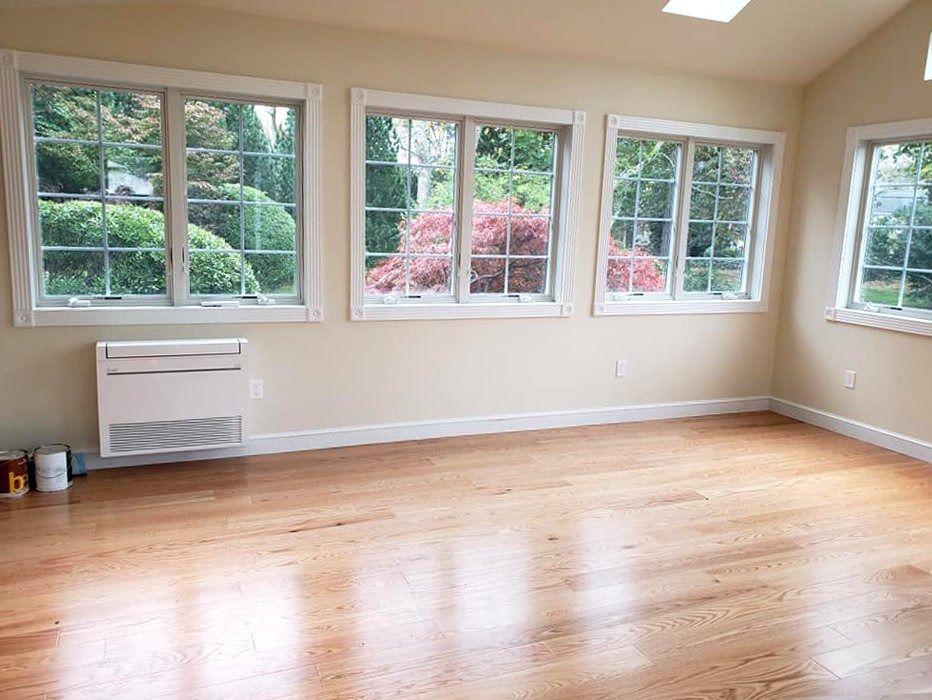 Empty room with hardwood floors, beige walls, and large windows overlooking greenery.