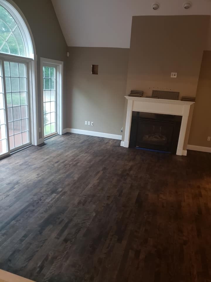 Empty living room with dark wood flooring, white fireplace, and large windows.