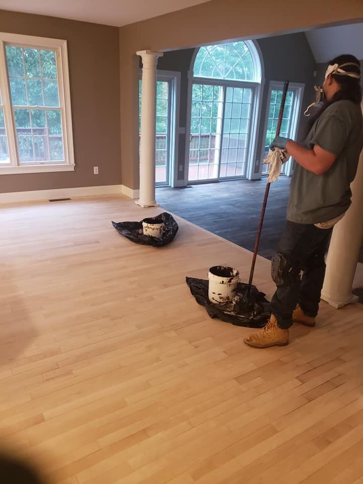 Person staining a light-colored wood floor in a spacious room with columns and windows. Paint supplies are on the floor.