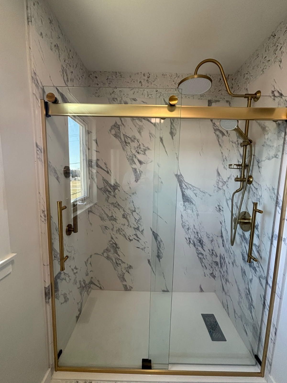 Gold-framed shower with marble-look walls, glass doors, and gold fixtures. White shower base and small window.