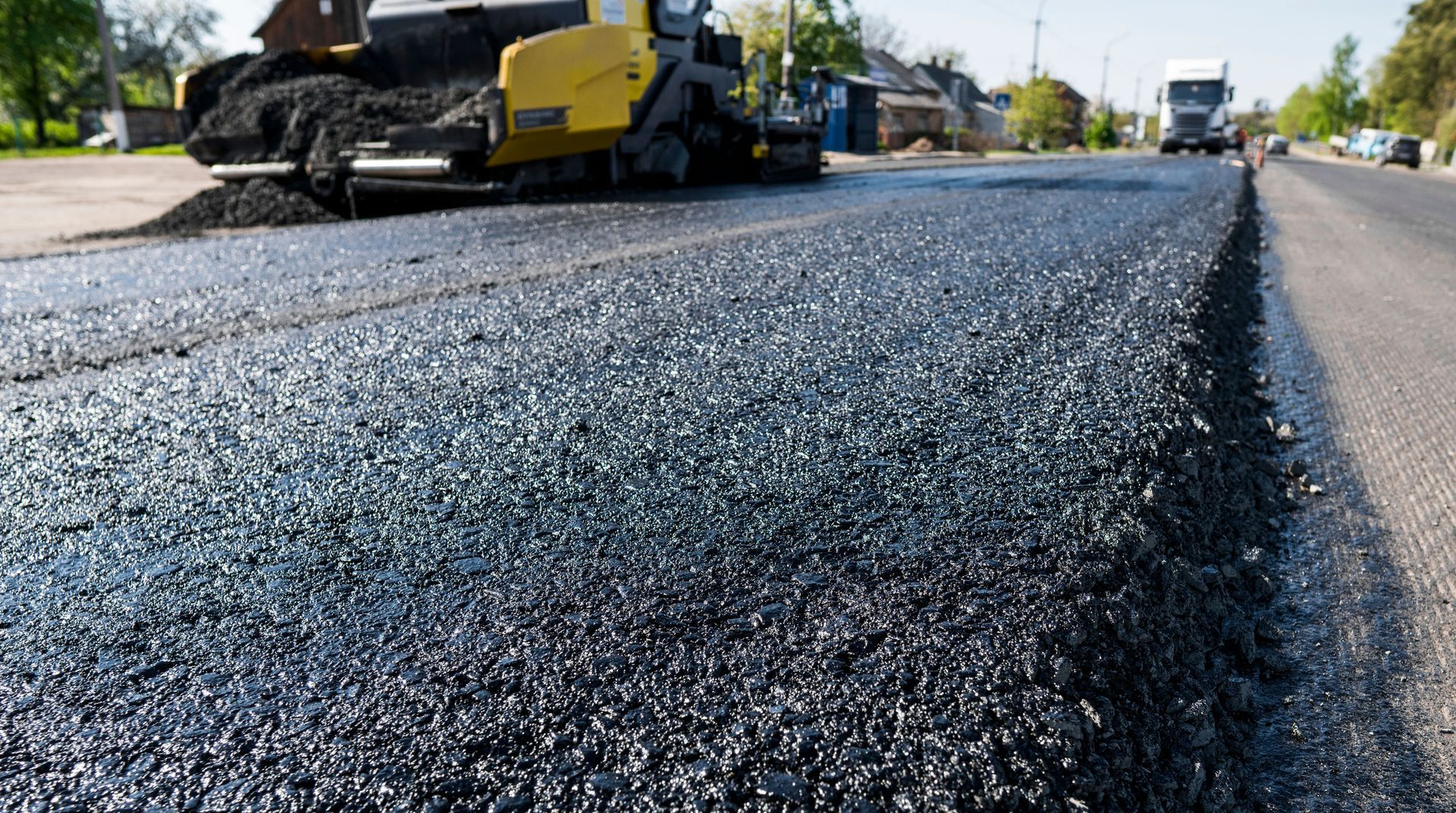 Asphalt paving a road.  A yellow paver machine spreads hot asphalt. A truck is visible in the distance.