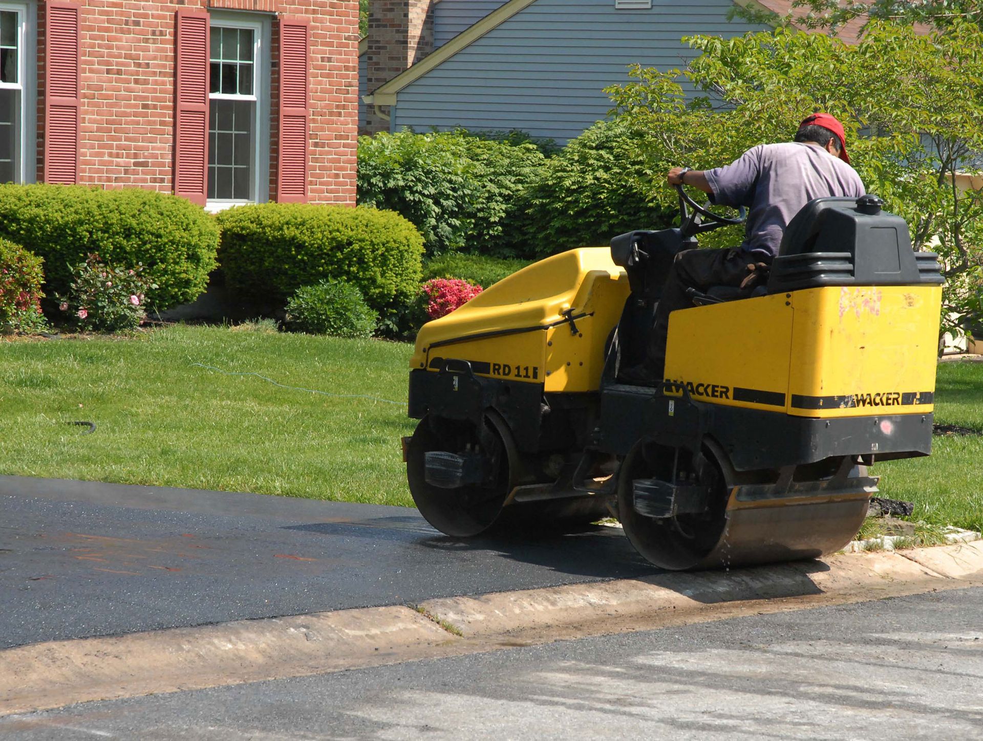 Yellow asphalt roller compacting fresh pavement beside a curb and green grass; a worker operates the machine.