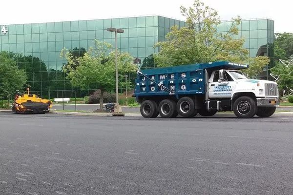 Asphalt paving: Dump truck and compactor on a paved lot in front of a modern glass building.