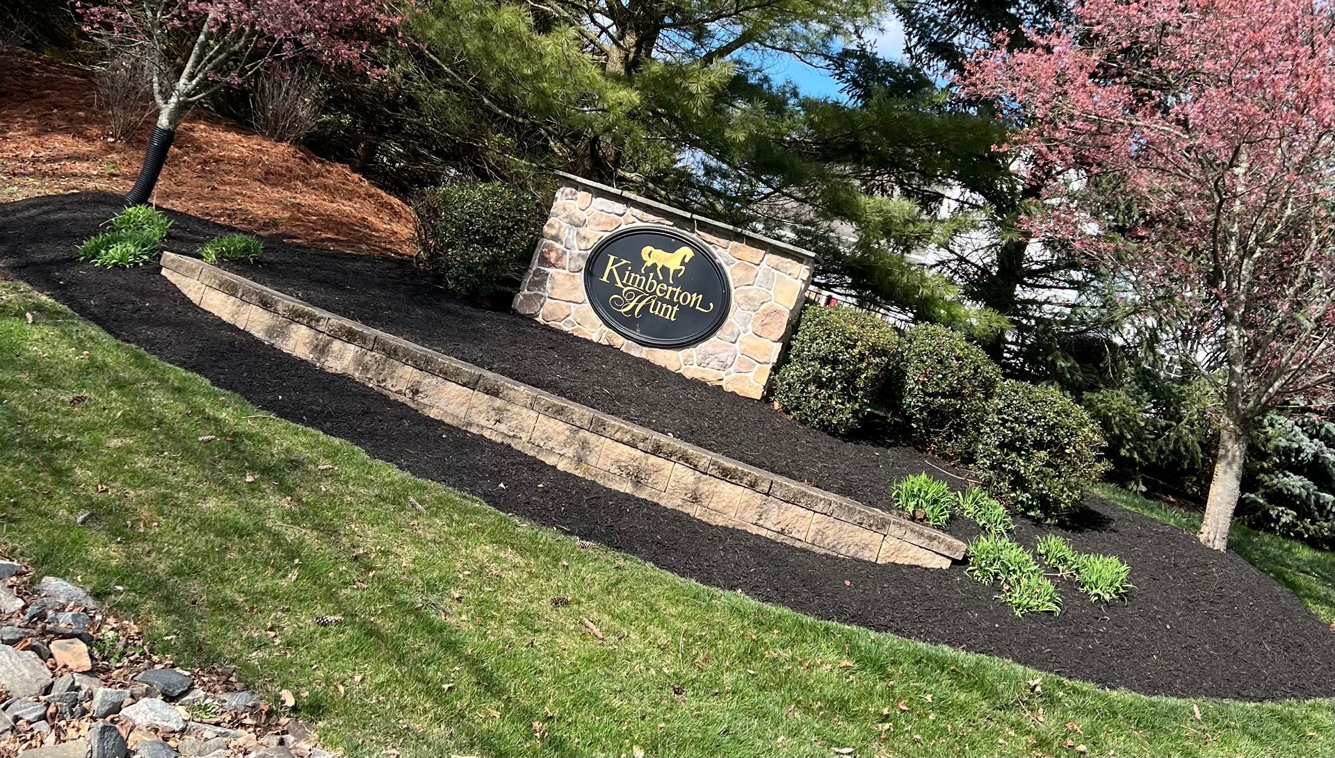 Stone sign in a landscaped garden with shrubs, grass, and blooming trees on a sunny day