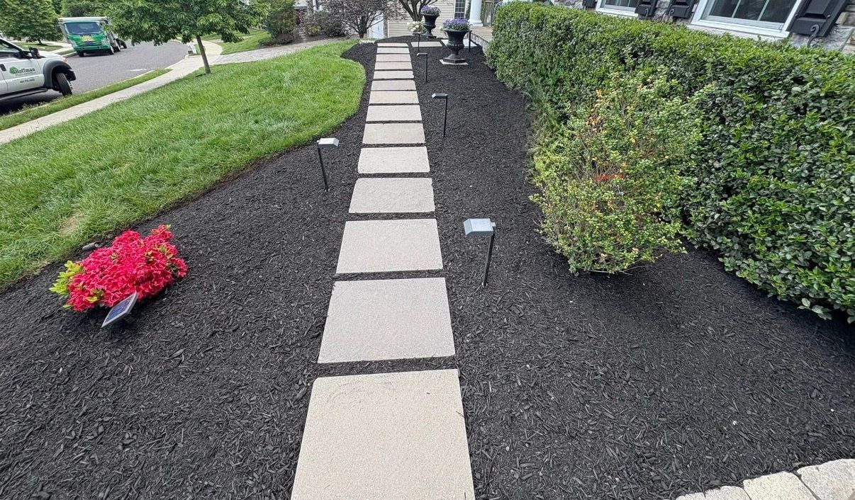 Stepping-stone walkway through black mulch with shrubs and a red flower pot beside a building