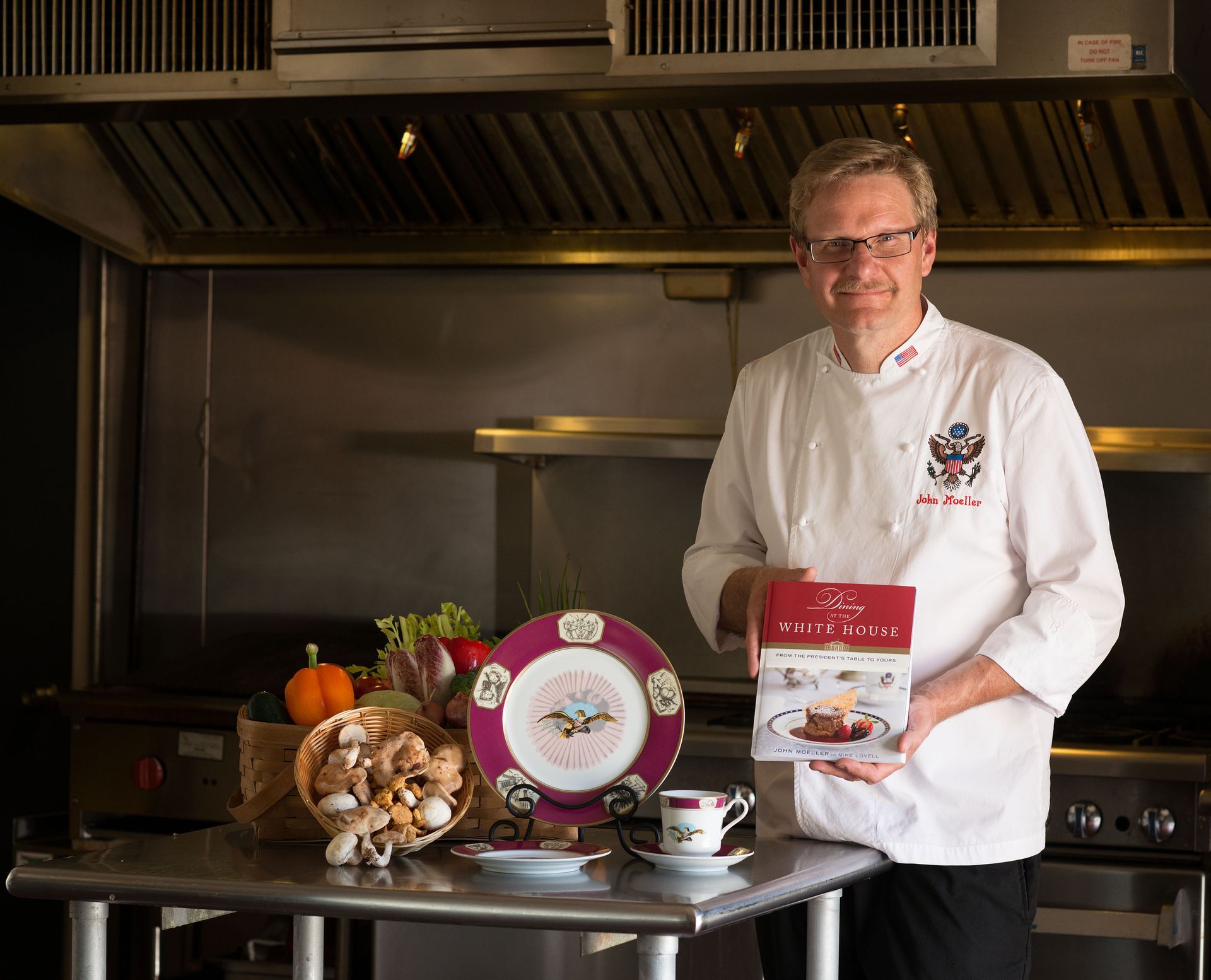 Chef John in a commercial kitchen displaying packaged food and a branded plate beside ingredients.