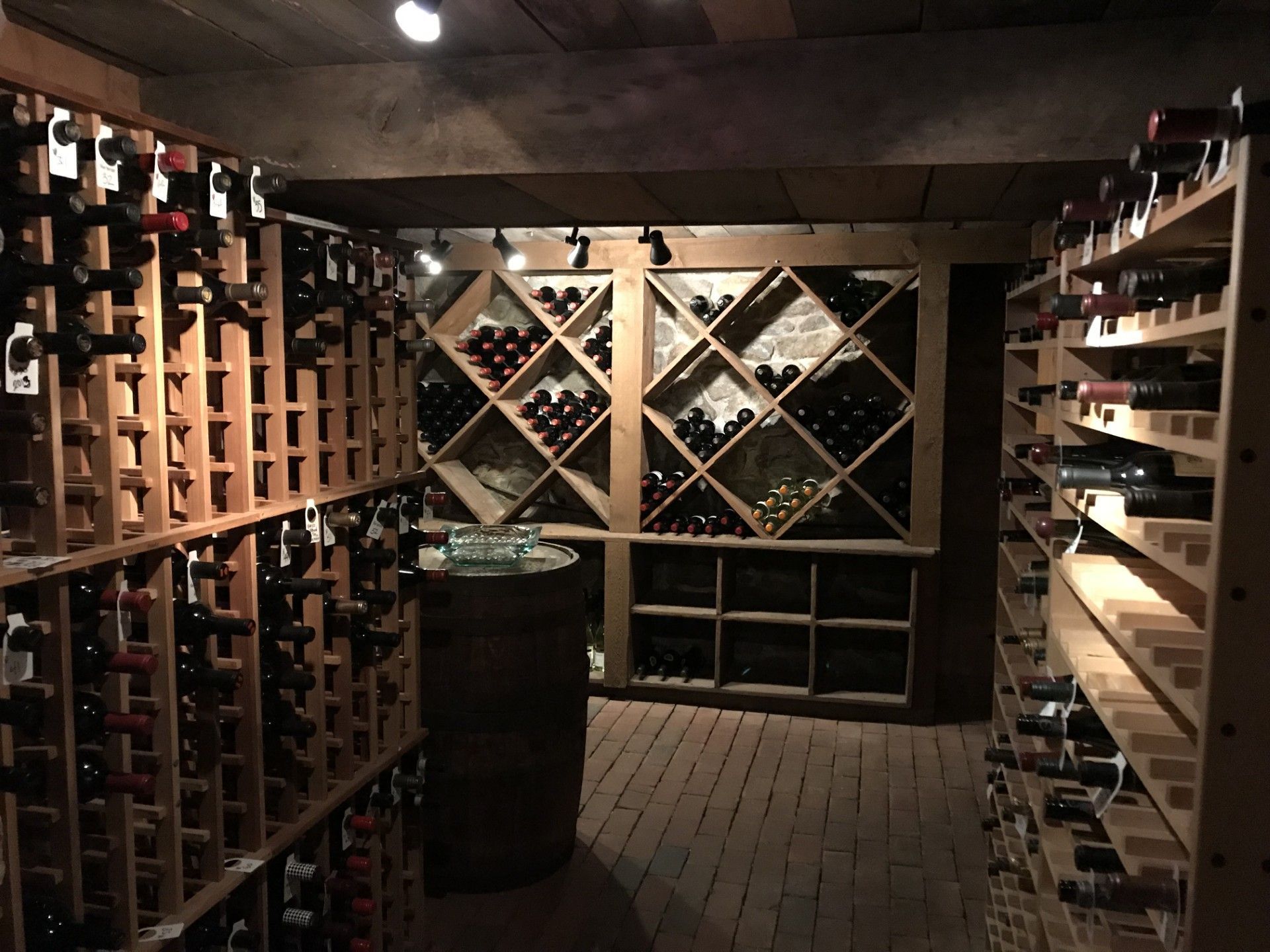 Wine cellar with wooden racks of bottles and a brick floor, dimly lit.