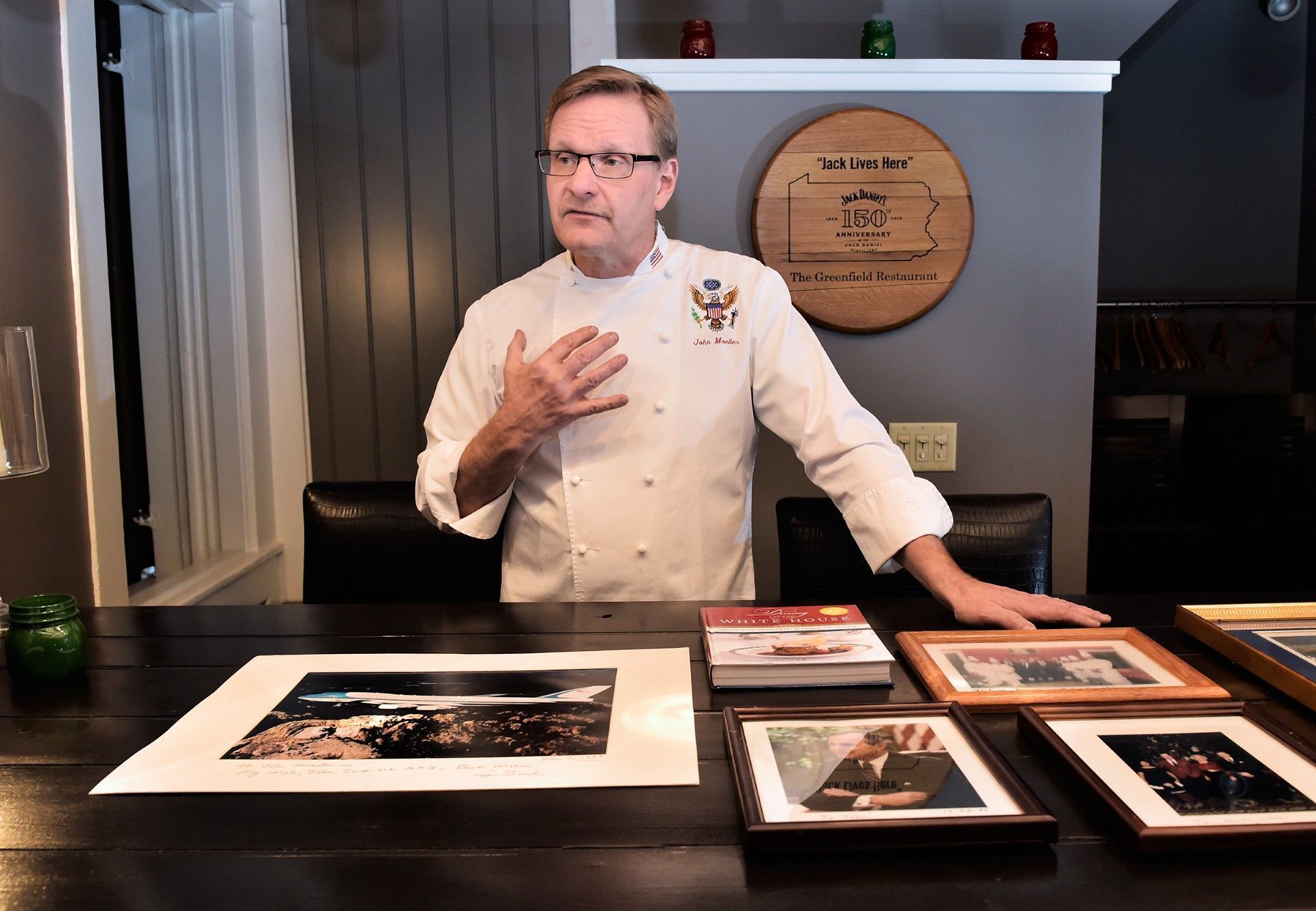 Chef John in a white coat presenting framed food photos on a table in a restaurant setting