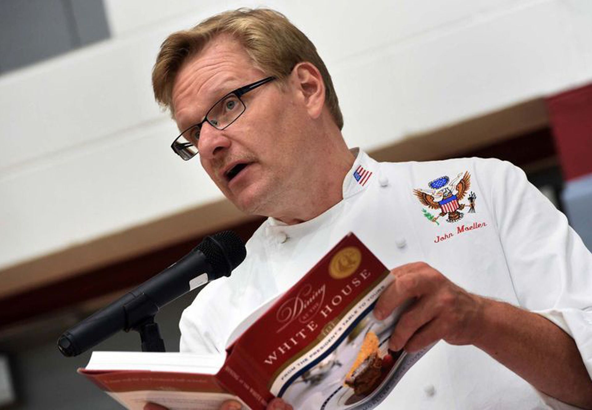 Chef John is reading aloud from a red book at a microphone indoors