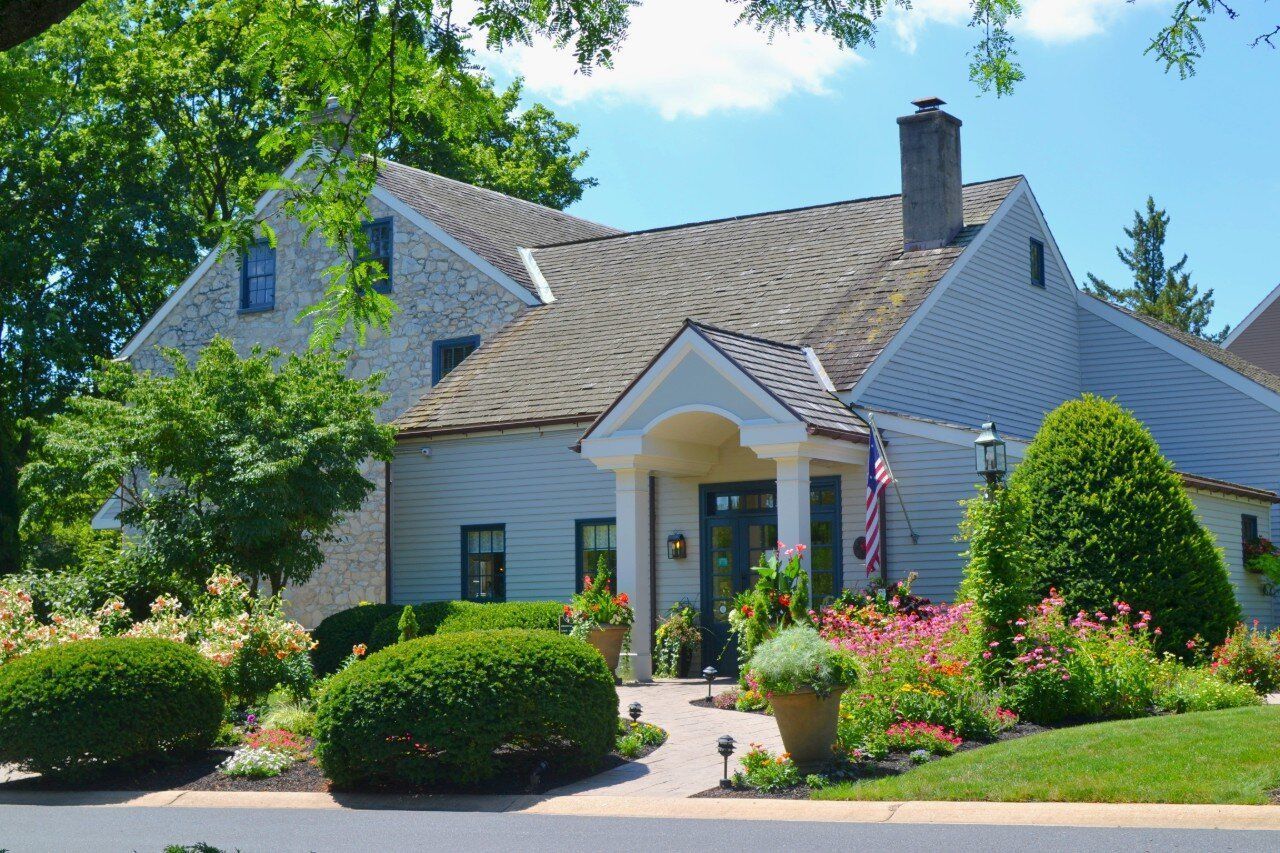 A white house with a brown roof is surrounded by bushes and flowers