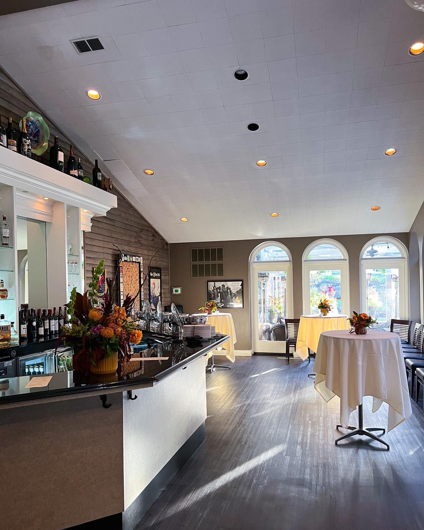 Bright banquet hall with bar counter, round tables, arched windows, and recessed ceiling lights