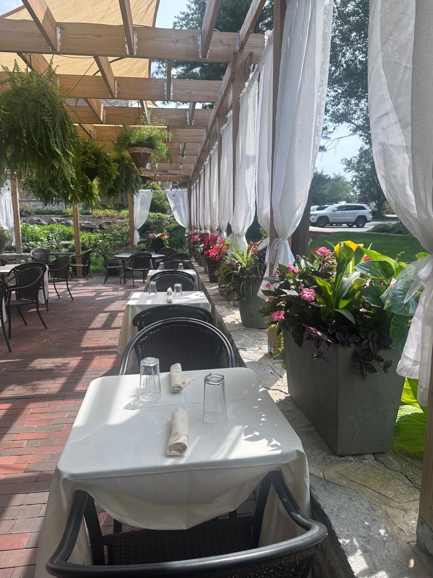 Outdoor café terrace with white tablecloth tables, wicker chairs, and potted plants under a wooden pergola.